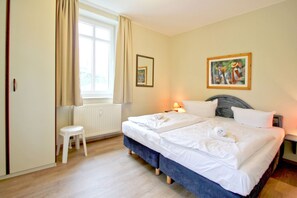 A hotel room with two beds, a window with curtains, a radiator, a small stool, and a painting on the wall. - Close to the beach, with balcony - Villa Strandmuschel FeWo 08 - Villa Strandmuschel (Göhren)