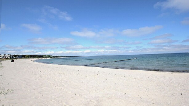 En bred sandstrand med stilla vatten, en klar himmel och några få byggnader i fjärran.