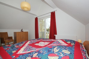 Une chambre à coucher avec un grand lit, une chaise en bois, une petite table, une fenêtre avec des rideaux rouges et un luminaire suspendu.