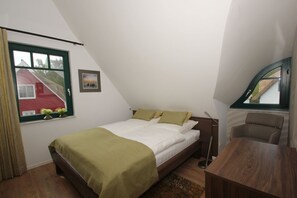 Un dormitorio con cama, escritorio y silla. Se ve una ventana con vista a un edificio rojo y follaje.