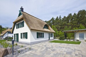 Una casa tradicional con techo de paja, paredes blancas y contraventanas verdes, rodeada de un jardín cuidado y un camino empedrado.