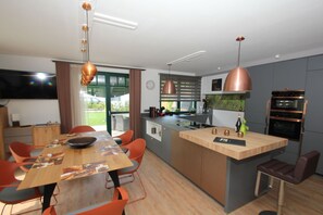 A modern kitchen with a central island, pendant lighting, and a dining area with orange chairs. - Ferienhaus Seehund - House in a great location with garden, terrace and private sauna (Glowe)