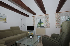 A living room with a sofa, armchair, glass coffee table, TV, and a radiator. - Waterfront house with terrace and garden - Ferienhaus Storchennest Haus L - (Wiek)