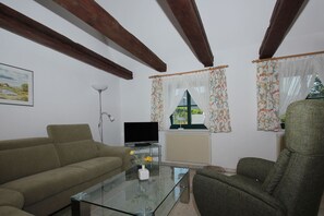 Ein Wohnzimmer mit Sofa, Sesseln, Couchtisch aus Glas, Fernseher und Heizkörper.