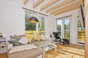 A modern living room with a sofa, a glass coffee table, and a black lounge chair. There are large windows and a wooden ceiling.