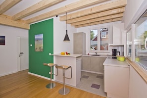 A compact kitchen with a green accent wall, wooden beams, a small bar area, and a window with a view of a house.