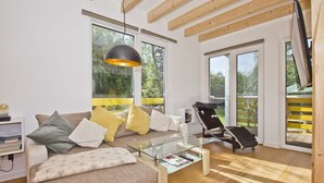 A modern living room with a sofa, a glass coffee table, and a black lounge chair. There are large windows and a wooden ceiling. - Vacation home Saga - close to the beach, modern house with own sauna, garden, terrace - (Wiek)