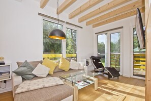 Ein modernes Wohnzimmer mit einer Couch, einem Glastisch und einem schwarzen Sessel. Große Fenster und eine Holzbalkendecke sind vorhanden.