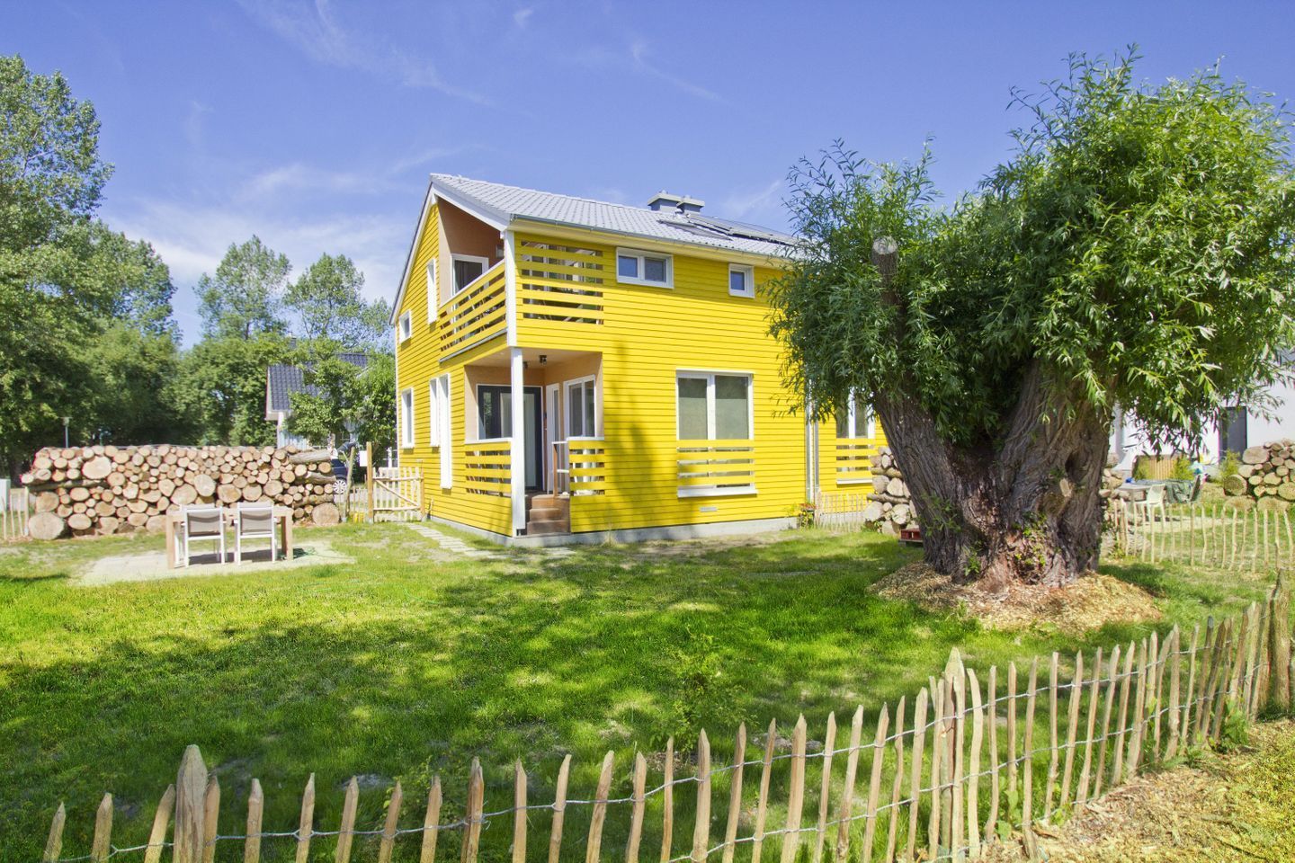 A yellow two-story house with a front porch, a wooden fence, and a large tree in the yard.