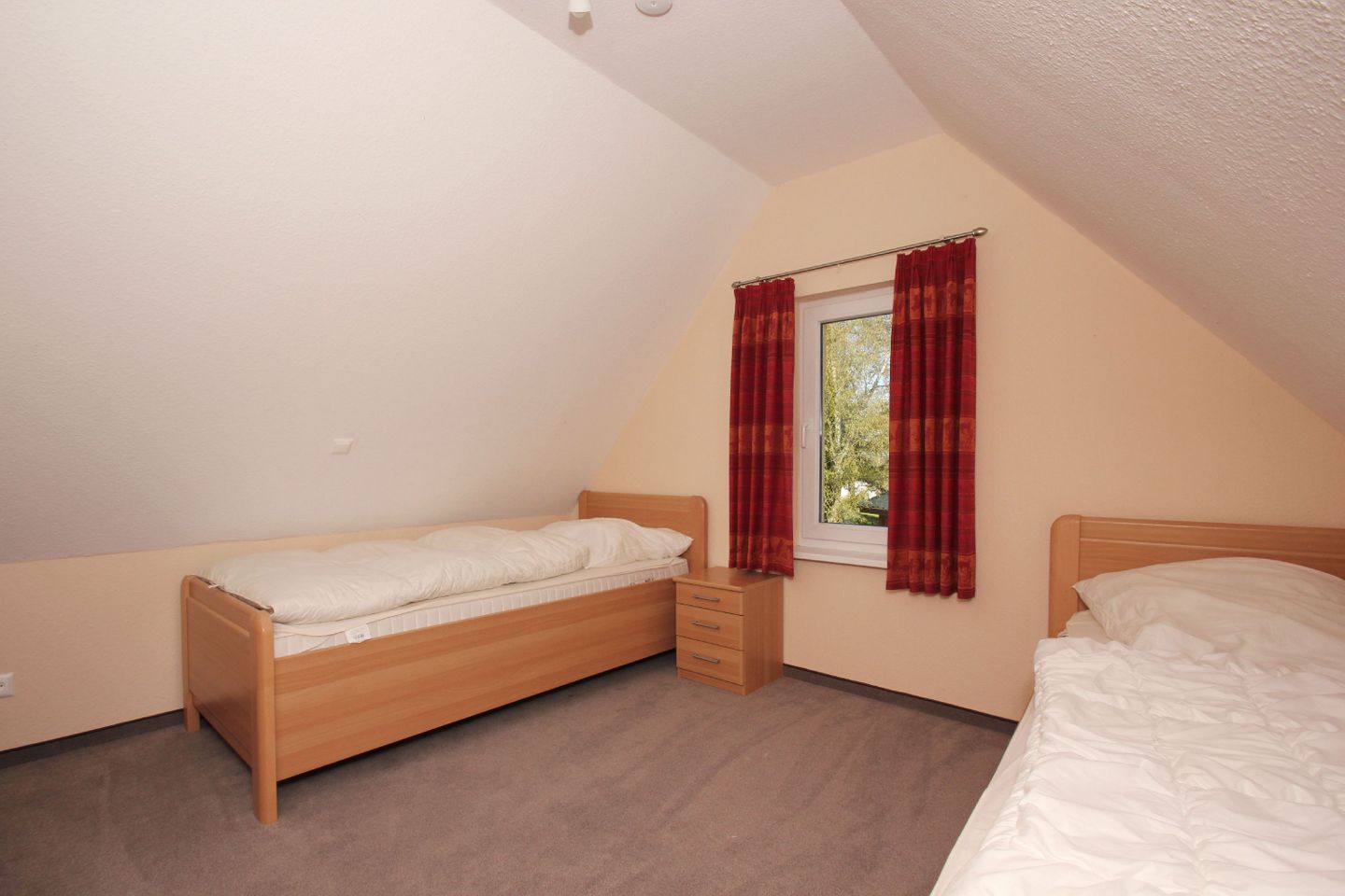 A small, single-room bedroom with two wooden beds, a window with red curtains, and a carpeted floor.