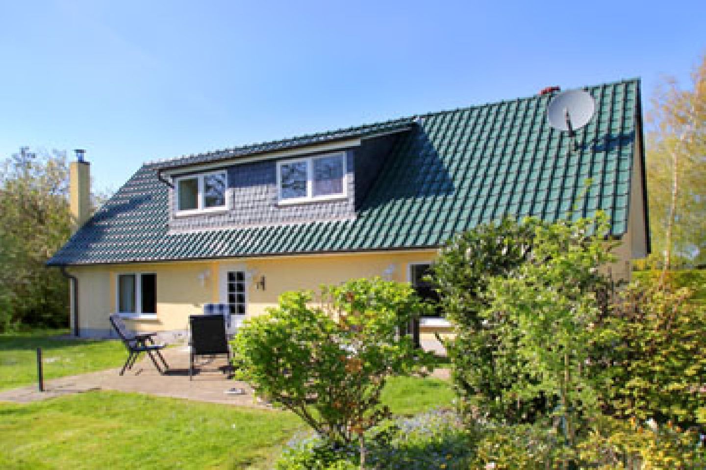 A two-story house with a green roof, a satellite dish, and a small garden area with a bench and a table.