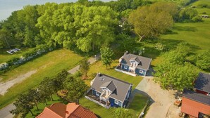 Un quartier résidentiel comprenant deux maisons principales, un grand arbre et une étendue d’eau en arrière-plan.