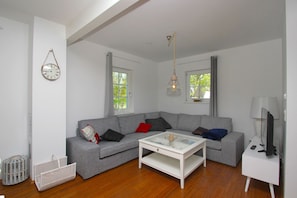 A modern living room with a grey sectional sofa, a white coffee table, and a television on a white stand. There are two windows with curtains, a hanging lamp, and a wall clock. - Freya Vacation Home - Cozy Swedish-style home near the beach with a view of the sea - (Wiek)