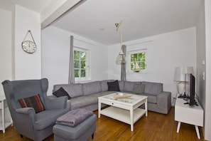 Un salon moderne avec un canapé gris, une table basse blanche et un fauteuil gris. Il y a une horloge au mur et une lampe sur une table d’appoint.