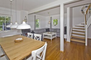 A spacious living area with a wooden dining table, white chairs, a gray sofa, and a staircase leading to an upper floor.