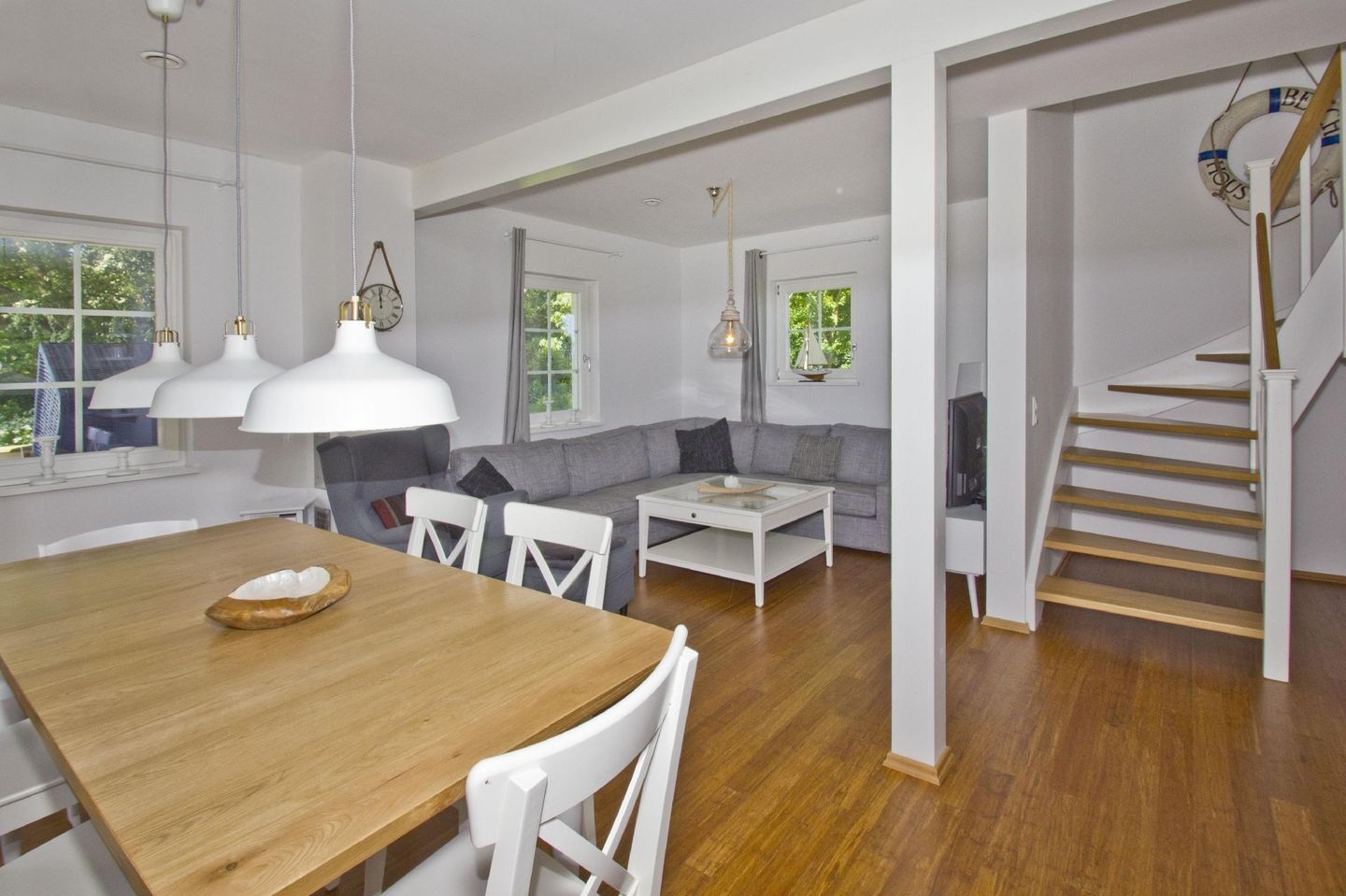 A spacious living area with a wooden dining table, white chairs, a gray sofa, and a staircase leading to an upper floor.