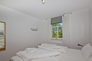 A bedroom with two beds, a window with curtains, a wall-mounted light, and a small mirror. - Apartment near the harbor with a balcony and ocean view - Hafenhäuser Breege FeWo 14 - (Breege)
