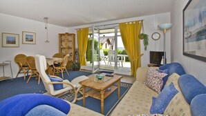 A living room with a sofa, chairs, a table, and a TV. There is a window with yellow curtains and a view of the outside.