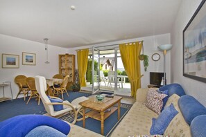 A living room with a sofa, chairs, a table, and a TV. There is a window with yellow curtains and a view of the outside.