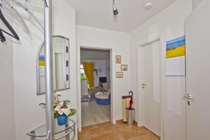 En hall med ett vitrinskåp, en spegel, en dörr med en kalender på och utsikt över ett vardagsrum med en gul gardin.