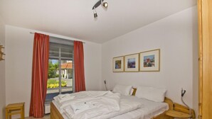 A bedroom with a large bed, a wooden headboard, a bedside table, a small stool, and a window with curtains.
