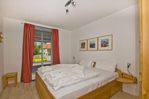 A bedroom with a large bed, a wooden headboard, a bedside table, a small stool, and a window with curtains. - Vacation apartment near the harbor with terrace and sea view - Hafenhäuser Breege FeWo 13 (Breege)