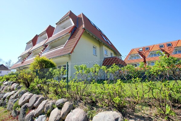 A multi-story residential building with a red-tiled roof, a balcony, and a garden area with a fence and rocks.