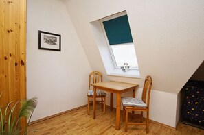 A small, cozy room with a sloped ceiling, a wooden table, two chairs, a framed picture on the wall, and a window with a view of the sky.