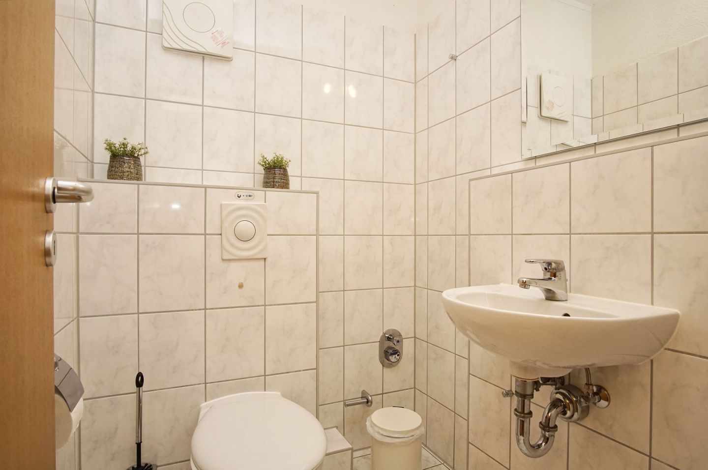 Ein Badezimmer mit einem weißen Waschbecken, einer Toilette und einem Händetrockner. An der Wand befinden sich zwei kleine Topfpflanzen.