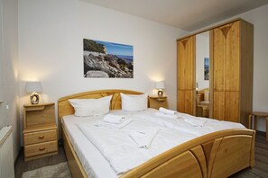 A bedroom with a wooden bed, white bedding, two bedside tables with lamps, a large wooden wardrobe, and a framed picture of a rocky coastline. - 2-room vacation apartment near the harbor with balcony - Hafenhäuser Breege FeWo 05 - (Breege)