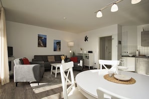Una sala moderna con un sofá gris, sillas blancas, una mesa de comedor y una cocina con armarios blancos y salpicadero de azulejos.