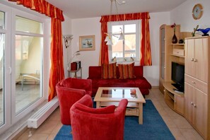 A living room with a red sofa, a red armchair, a glass coffee table, a TV stand, and a window with curtains. - Apartment near the harbor with balcony and sea view - Hafenhäuser Wiek 15 - (Wiek)
