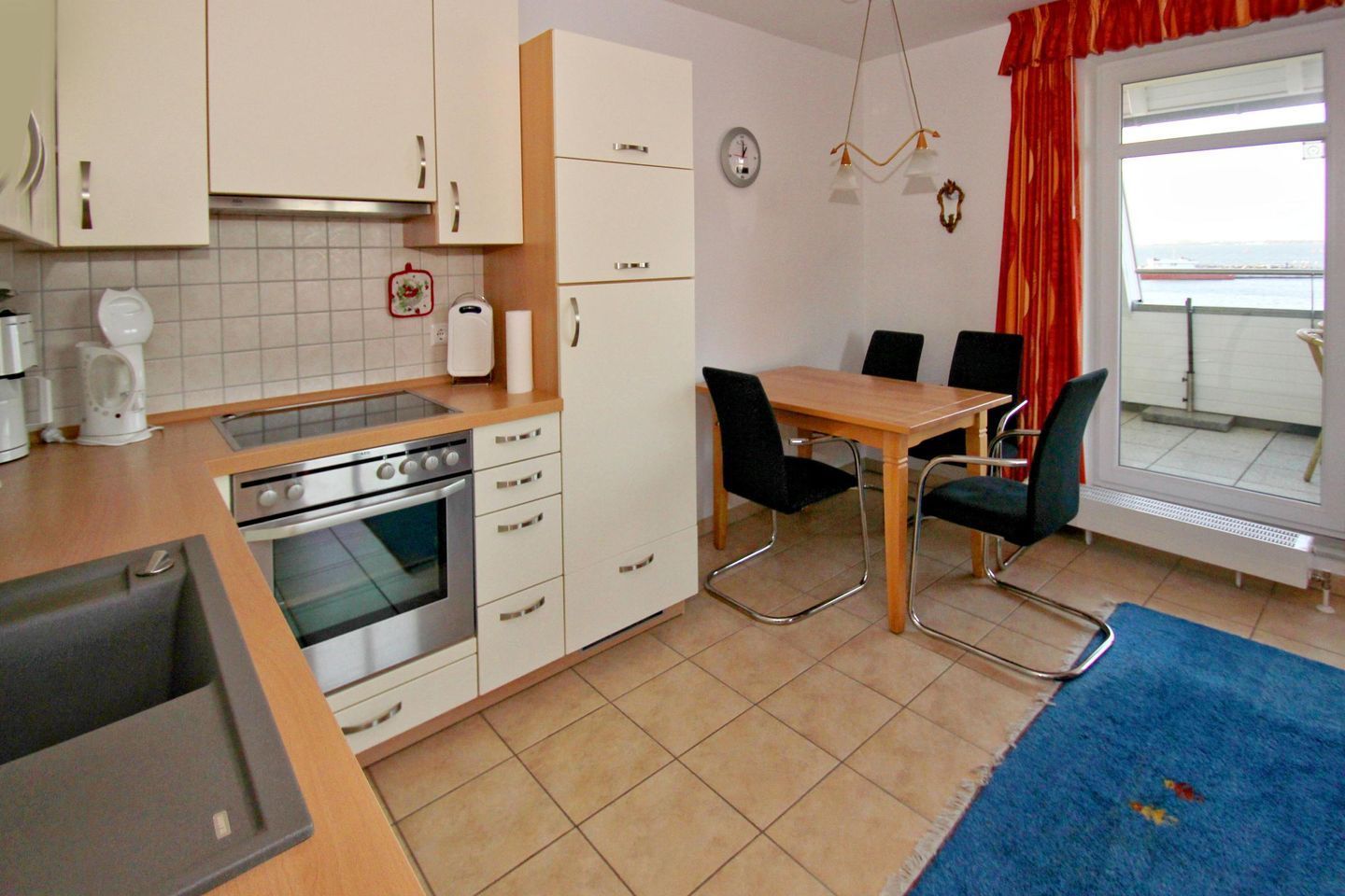A compact kitchen with a stove, oven, and white cabinets. There is a dining table with chairs and a view of the sea through a window.