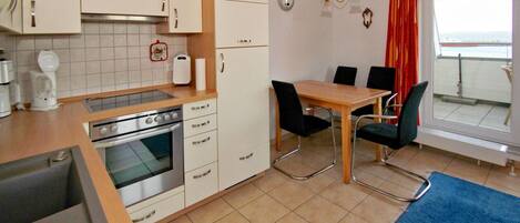 A compact kitchen with a stove, oven, and white cabinets. There is a dining table with chairs and a view of the sea through a window.
