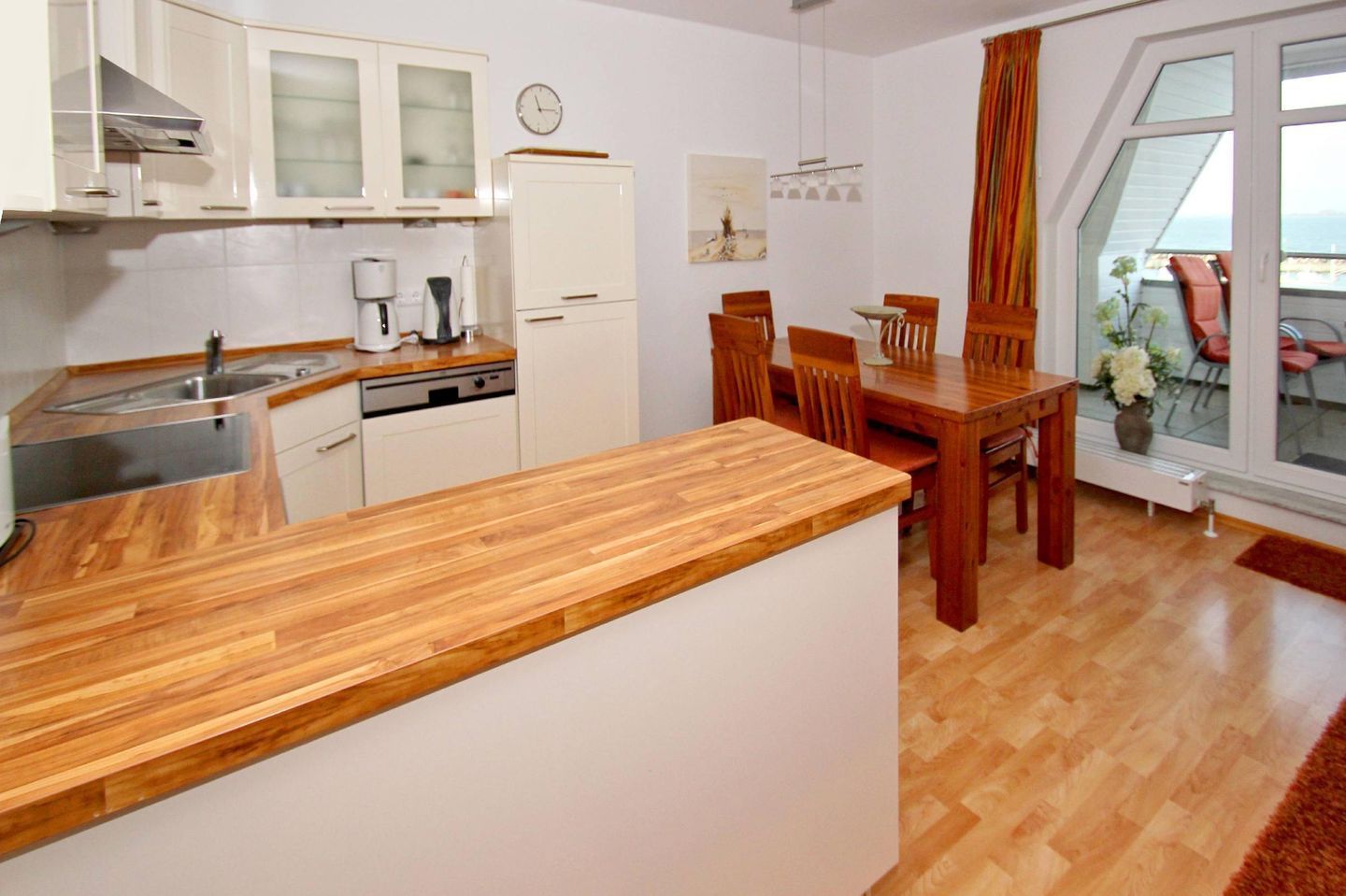 Una cocina compacta con armarios blancos, encimera de madera y una zona de comedor con sillas y mesa de madera.