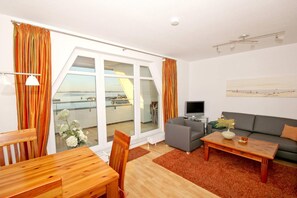A modern living room with a sofa, coffee table, and TV. Large windows offer a view of the sea.