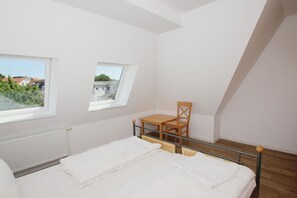 Ein kleiner, heller Raum mit einem Bett, einem Holzhocker, einem kleinen Tisch und einem Fenster.