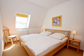 Un dormitorio con una cama individual, una silla, una mesita de noche, una lámpara, un ventanal y un cuadro en la pared.