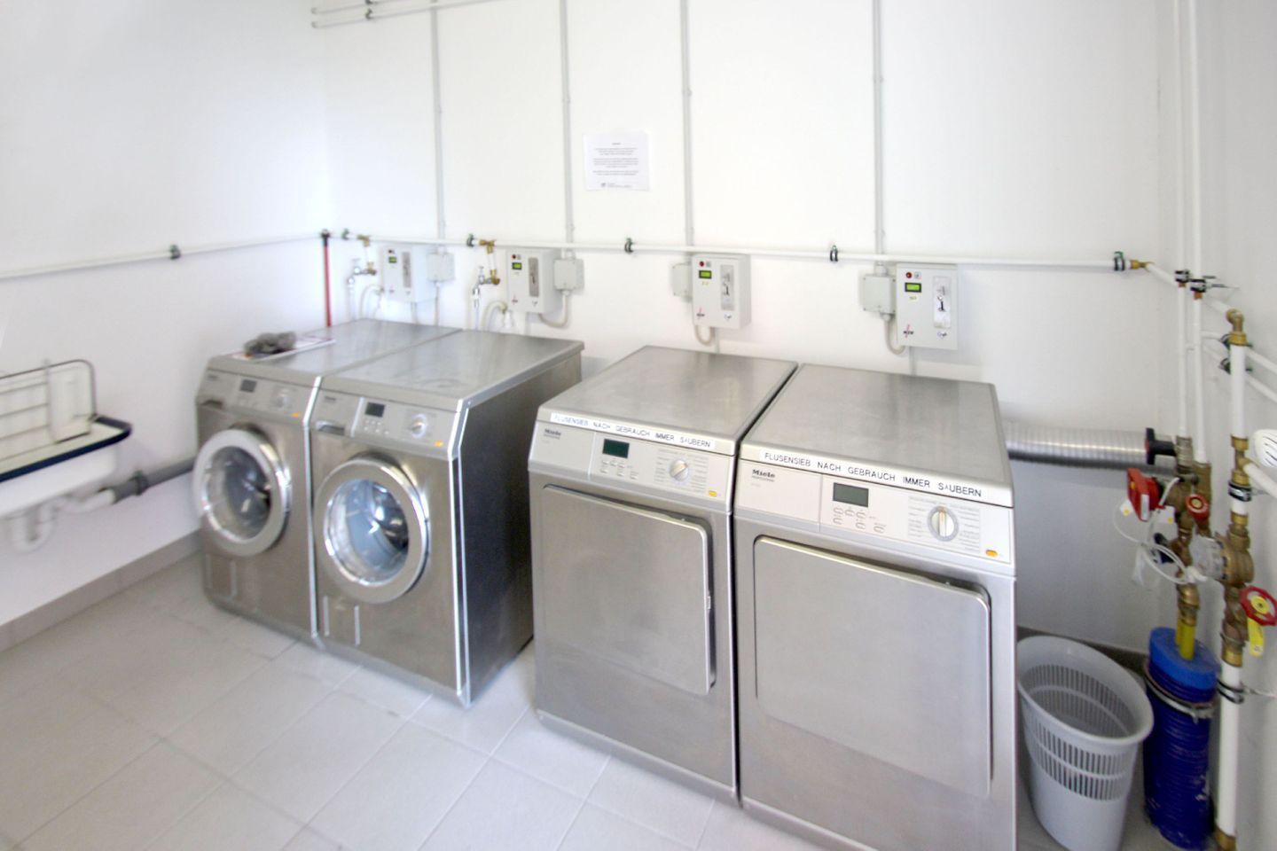 A laundry room with two front-loading washing machines and a dryer, equipped with various control panels and hoses.