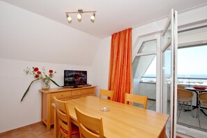 A modern dining area with a wooden table and chairs, a TV on a wooden cabinet, and a view of the sea through large windows. - Apartment near the harbor with balcony and sea view - Hafenhäuser Wiek FeWo 04 - (Wiek)
