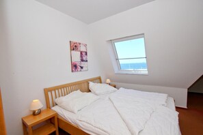 A bedroom with a wooden bed, white bedding, a nightstand with a lamp, a window with a view, and a wall-mounted artwork. - Apartment near the harbor with balcony and sea view - Hafenhäuser Wiek 06 - (Wiek)