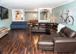 A living room with a sectional sofa, a blue armchair, and a dining area with wooden chairs. - Central Lake George Escape – Balcony Views, Walk to Everything (Lake George)