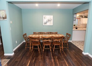 A dining area with a wooden table and chairs, light blue walls, and a kitchen in the background.