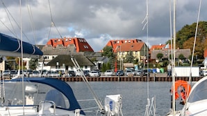 Un port de plaisance avec des bateaux amarrés, une jetée où sont garées des voitures, et des bâtiments aux toits rouges en arrière-plan.