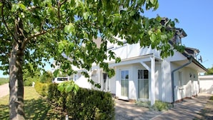 Une maison blanche à deux étages, dotée d’un porche abrité, d’une allée et d’une haie devant.