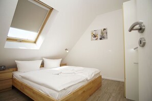 A bedroom with a sloped ceiling, a bed with white linens, a wooden headboard, a nightstand, a wall-mounted light, and a door with a keyhole. - Cozy vacation apartment near the harbor - FeWos am Hochzeitsberg FeWo 10 - FeWos (Breege)