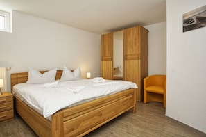 A neatly made bed with white linens and pillows, a wooden wardrobe, a small orange chair, and a bedside table with a lamp. - Cozy vacation apartment near the harbor - FeWos am Hochzeitsberg FeWo 17 - FeWos (Breege)