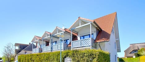 A row of white houses with red roofs and blue balconies, surrounded by a white picket fence and greenery.