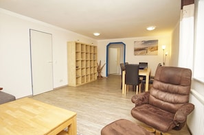 A living room with a brown leather sofa, a wooden coffee table, a dining table with chairs, and a bookshelf.