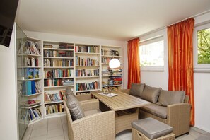 A cozy reading nook with wicker furniture, a wooden table, and a large bookshelf filled with books.
